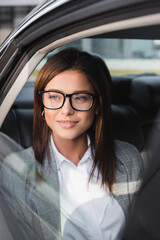 stylish, businesswoman in eyeglasses looking out from open window in car on blurred foreground