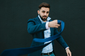 Young man, with jacket and shirt, looking like an entrepreneur, moving his tie. In a black wall of the street background. Work and young entrepreneurs concept