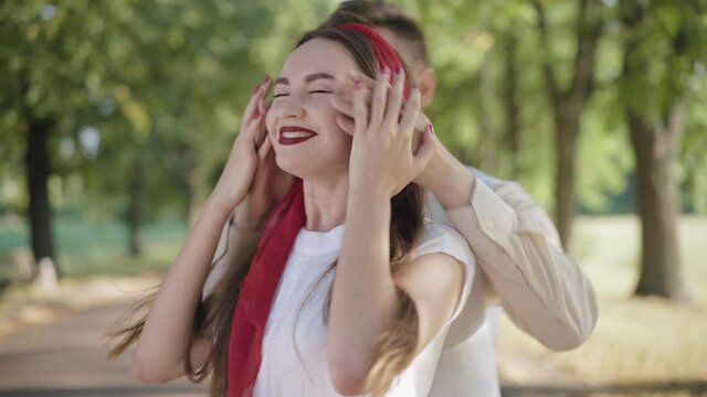 Cheerful young man closing eyes of beautiful young woman waiting for a date and hugging adorable partner. portrait of happy Caucasian boyfriend and girlfriend meeting in sunny summer park.