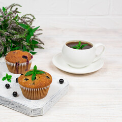 Cupcakes with black currants on a white wooden cutting board against the background of a cup and mint flowers.