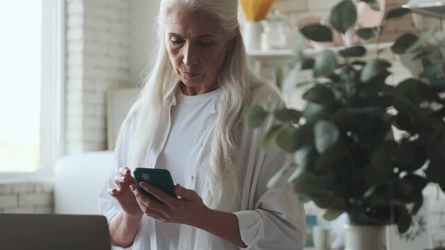Concentrated senior woman using laptop computer and mobile phone at the kitchen indoors at home