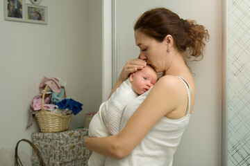 Mom gently kisses her newborn baby at home against the backdrop of a basket of laundry. The mother gently holds the child in her arms. Household chores and concerns of a lonely young mother. Copyspace