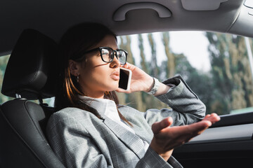  businesswoman in eyeglasses gesturing while talking on smartphone in car on blurred foreground