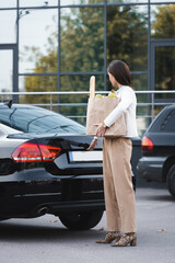  woman opening trunk of black car while holding shopping bag with food