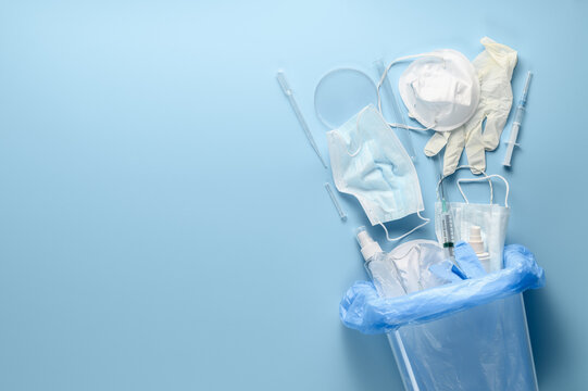 Coronavirus protection equipment in medical waste bin. Used disposable medical face masks, latex medical gloves, syringes and test tubes on pastel blue background. Flat lay, place for text