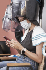 one hispanic young adult woman sitting in a hair dryer hood beauty salon or spa using protective mask and using smartphone during coronavirus covid-19 pandemic
