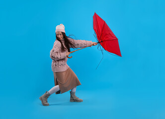 Young woman with umbrella caught in gust of wind on light blue background