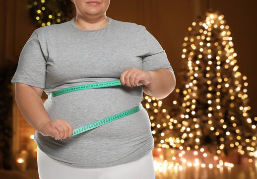 Overweight Woman Measuring Her Waist In Room With Christmas Tree After Holidays, Closeup