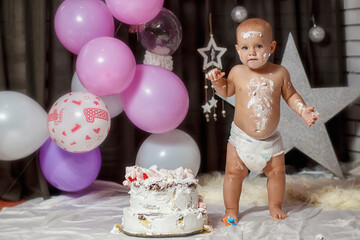 A little girl is standing in diapers and eating a cake on her first birthday. The baby eats a delicious cake with his hands. Happy Birthday.