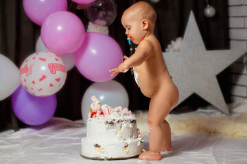 The first birthday of a baby standing naked near a birthday cake. Little girl stands near a cake in her room decorated with balloons. Happy birthday card