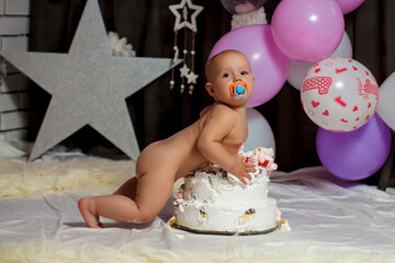 A little girl with a pacifier in her mouth lies naked on a birthday cake on her first birthday. Infant has fun in his baby room decorated with balloons. Happy birthday card