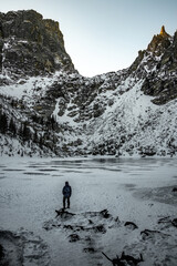 View of a person standing in front of Emerald Lake in Rocky Mountain National Park in Colorado during the winter. The lake is frozen over and the mountain range is covered in snow.