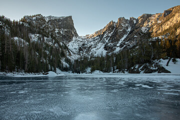 A view of Dream Lake in Rocky Mountain National Park with snow covered peaks surrounded by a forest of trees, snow, and ice. The lake is frozen and has distinct texture.