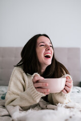 portrait of a white woman with freckles and dark hair laughing out loud lying on the bed while holding a bowl