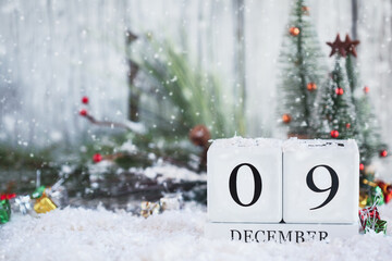 White wood calendar blocks with the date December 9th and Christmas decorations with snow. Selective focus with blurred background. 