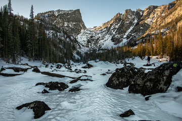 A view of Dream Lake in Rocky Mountain National Park with snow covered peaks surrounded by a forest of trees, snow, and ice. Shot is taken with amongst rocks and a frozen stream.