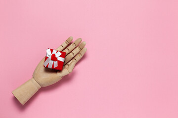 Wooden hand holds small red gift box with white bow and little heart on pink background, copy space, flat lay. March 8, February 14, birthday, Valentine's, Mother's, Women's day celebration concept