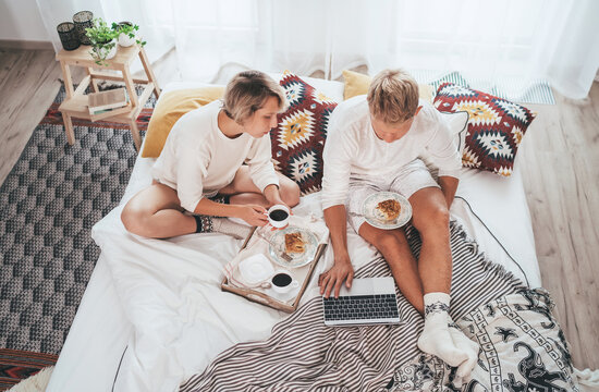 Teens couple in pajamas in cozy bed browsing internet together using a laptop and having a morning coffee with apple pie dessert. Couples relations and internet technology top view concept image.