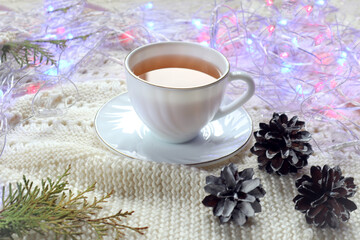 Obraz na płótnie Canvas A Cup of hot tea with pine cones and a multicolored garland on a white knitted sweater, side view, close - up-the concept of the arrival of pleasant holiday days