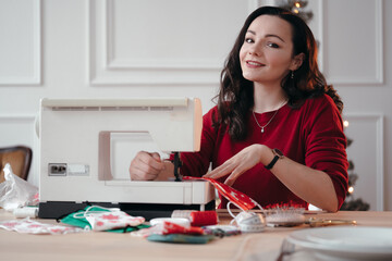 Attractive Caucasian girl sits at a sewing machine against the background of a Christmas tree. The seamstress makes homemade Christmas gifts with her own hands