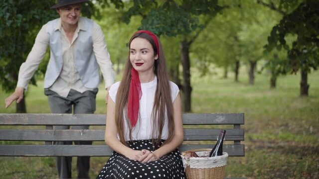 Elegant Caucasian boyfriend walking from the background to beautiful girlfriend sitting on bench waiting for a date. Confident handsome man joining slim charming woman and giving compliments.