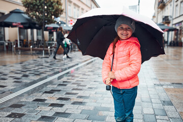 Little smiling happy girl holding big umbrella walking in a downtown on rainy gloomy autumn day