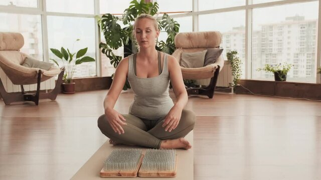 Full shot of woman sitting in lotus pose placing hands on sharp nails of sadhu board then starting meditation with her eyes closed in spacious yoga studio