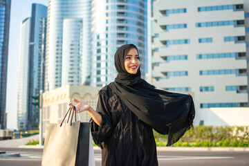 Beautiful young arab muslim woman in abaya clothes holding shopping bags and walking on the city street. Shopping time. Modern skyscrapers in the background.