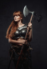 Obraz na płótnie Canvas Dressed in dark armour female scandinavian barbarian with brown hairs holding two handed axe in dark background.