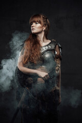 Fototapeta premium Portrait of warlike female fighter from north with long brown hairs in dark armour with two handed axe in dark smokey background.
