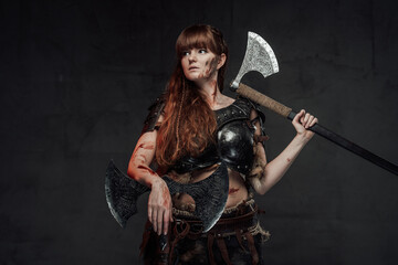 Obraz na płótnie Canvas Seductive and strong scandinavian woman fighter wielding two axes and dressed in dark light armour poses in dark background.