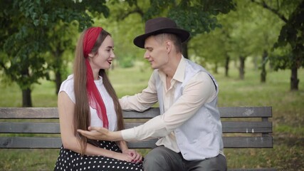 Elegant young Caucasian couple sitting on bench and chatting in summer park. Handsome man taking off hat and kissing woman behind it. Happy boyfriend and girlfriend dating outdoors. - Powered by Adobe
