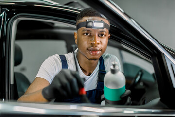 Close up of African man, car detailing service worker, cleaning car interior with soft brush, coating leather surface with professional anti dust and scratch liquid. Focus on man, looking at camera