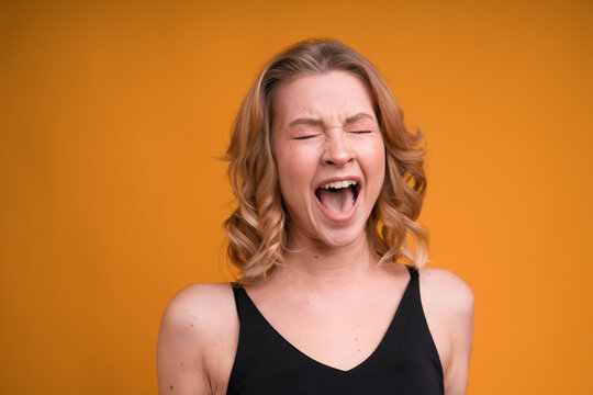 A portrait of a pretty Caucasian girl screaming loudly with her eyes closed and mouth opened, isolated on orange background