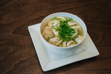 Dumplings with meat in broth with herbs in a white plate on a dark background. Russian folk cuisine. White bowl on a wooden table.
