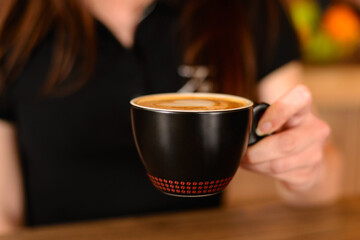 Close up of female hand holding a black cup of coffee with beautiful heart-shaped foam mid air over brown rustic wooden table. Focus on hands and cup.