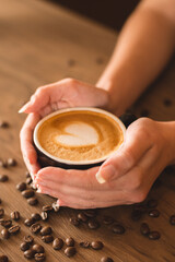 Close up vertical photo of female hands holding a black cup of coffee with beautiful heart-shaped foam on the top of brown wooden table with scattered coffee beans. Focus on hands and cup.
