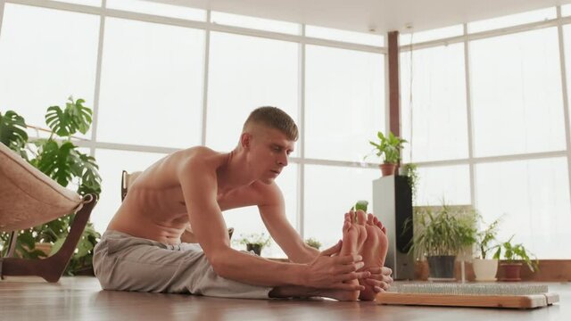 Full shot of professional yogi sitting on floor alone stretching and massaging his feet after meditating on sadhu nail board in bright yoga studio