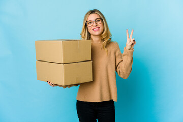 Young blonde caucasian woman holding boxes to move showing number two with fingers.