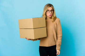 Young blonde caucasian woman holding boxes to move shrugs shoulders and open eyes confused.