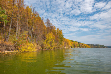 Autumn forest trees are reflected in the river water of the panoramic landscape. Blue sky with clouds.