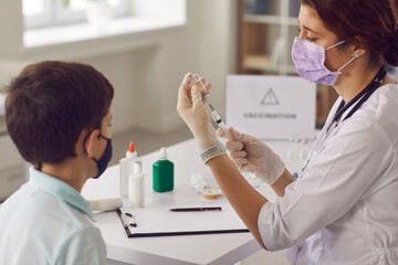 Little boy in face mask looking at syringe needle while nurse is getting vaccine ready