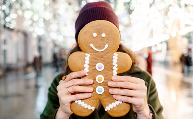Young beautiful girl, woman with huge funny gingerbread man. Christmas shopping. Walking on market street in big city decorated with garlands, lights.Stylish look, wool coat, hat.New year celebration
