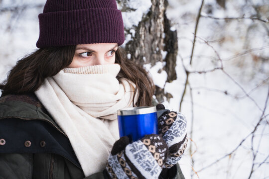 Young girl, woman in knitted mittens drinks tea, drinks coffee from thermo mug. Walking in beautiful winter forest covered with snow. Magnificent nature. Fashionable image, clothes, parka, hat, scarf