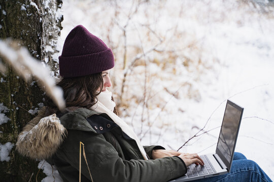 Young girl, woman working in laptop. Walking in beautiful winter forest among trees, firs, covered with snow. Magnificent nature and views. Fashionable image, clothes, parka, hat, mittens, blue jeans