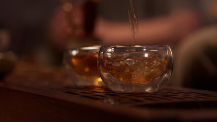 Cozy atmosphere of a tea room in a Chinese province. A stream of fragrant tea from a porcelain teapot fills two glass bowls. Close-up, warm light. Slowmo.