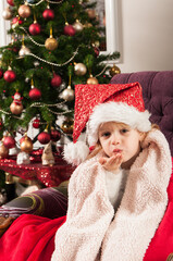Beautiful little girl with santa claus hat wrapped into red blanket, sitting beside christmass tree and sending kisses