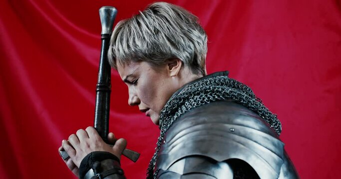 Side view of unhappy woman in metal medieval armor leaning on sword and worshipping before battle while impersonating Joan of Arc against waving red flag. Sad female warrior against red banner