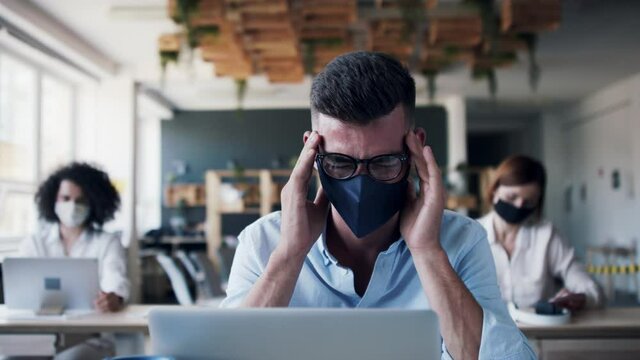 Portrait of young businesspeople with face masks working indoors in office. Young man having headache.