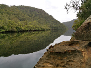 Fototapeta premium Beautiful view of a creek with reflections of cloudy sky, mountains and trees on water, Crosslands Reserve, Berowra Valley National Park, New South Wales, Australia 